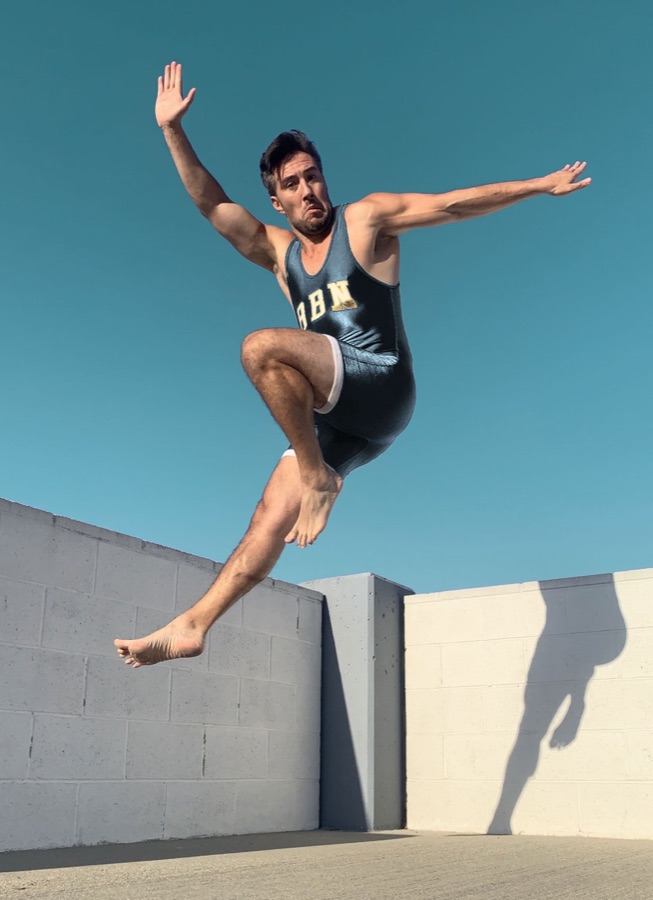 Max mid-jump in a vintage athletic singlet against a blue sky