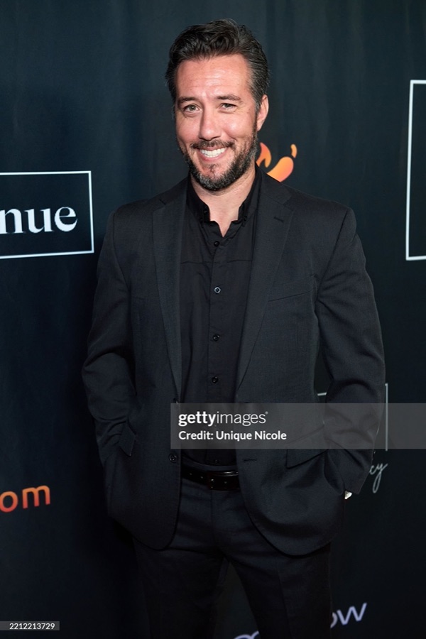 Max in an all-black suit on a step-and-repeat