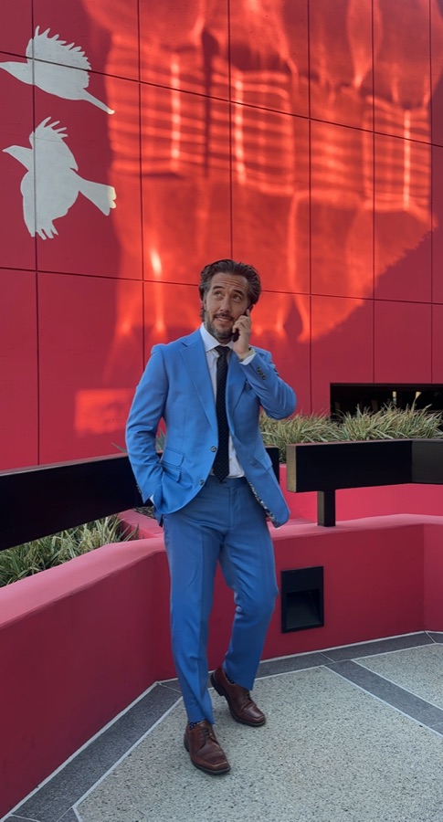 Max in a light blue suit on the phone, red wall backdrop, leading-man framing